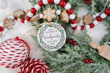 Load image into Gallery viewer, Personalized Ceramic Christmas ornament First Christmas as Grandparents with the baby's name and year. Adorned with a green wreath and red berries. The ornament is displayed on a table with Christmas Decorations.
