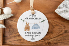 Load image into Gallery viewer, Personalized ornament for first grandchild with a brown pair of pants and blue sweater on a clothes line. Displaying the baby's last name and due year. The product is displayed on a wooden tray with decorative elements.