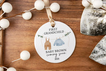 Load image into Gallery viewer, Personalized ornament for first grandchild with a brown pair of pants and blue sweater on a clothes line. Displaying the baby's last name and due year. The product is displayed on a wooden tray with decorative elements.