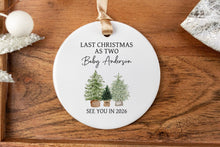 Load image into Gallery viewer, Personalized Christmas ornament made from white ceramic that says last Christmas as two with the baby's last name with two big trees and one little tree. the bottom says see you in the year the baby is to be born. The ornament is displayed on a wooden tray.