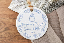Load image into Gallery viewer, Round white christmas ornament with the year we got engaged floral design, resting on a wooden surface with decorative elements.