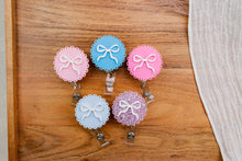 Load image into Gallery viewer, Five scalloped bow badge reels in pastel pink, blue, and glitter finishes on a wooden surface.