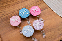 Load image into Gallery viewer, Pink, blue, and glitter scalloped bow badge reels arranged on a wooden tray.