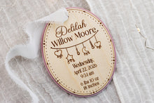 Load image into Gallery viewer, Angled view of an oval wooden baby keepsake ornament with pink acrylic backing, engraved birth details, and white ribbon.