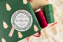 Load image into Gallery viewer, Personalized Ceramic Christmas ornament First Christmas as Grandparents with the baby's name and year. Adorned with a green wreath and red berries. The ornament is displayed on a table with Christmas Decorations. 
