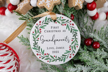 Load image into Gallery viewer, Personalized Ceramic Christmas ornament First Christmas as Grandparents with the baby's name and year. Adorned with a green wreath and red berries. The ornament is displayed on a table with Christmas Decorations. 
