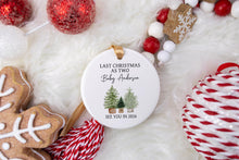 Load image into Gallery viewer, Personalized Christmas ornament made from white ceramic that says last Christmas as two with the baby's last name with two big trees and one little tree. the bottom says see you in the year the baby is to be born. The ornament is displayed on a white fur background with gingerbread deocrations and whtie and red ornaments. 
