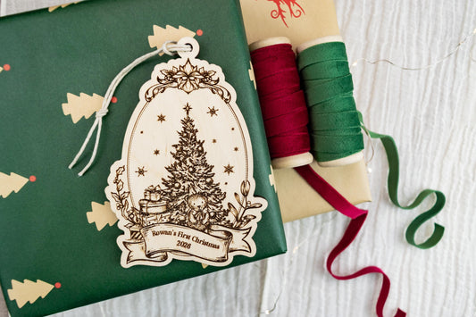 Vintage-style wooden Baby's First Christmas ornament with engraved Christmas tree, presents, stars, floral border, and personalized banner.
