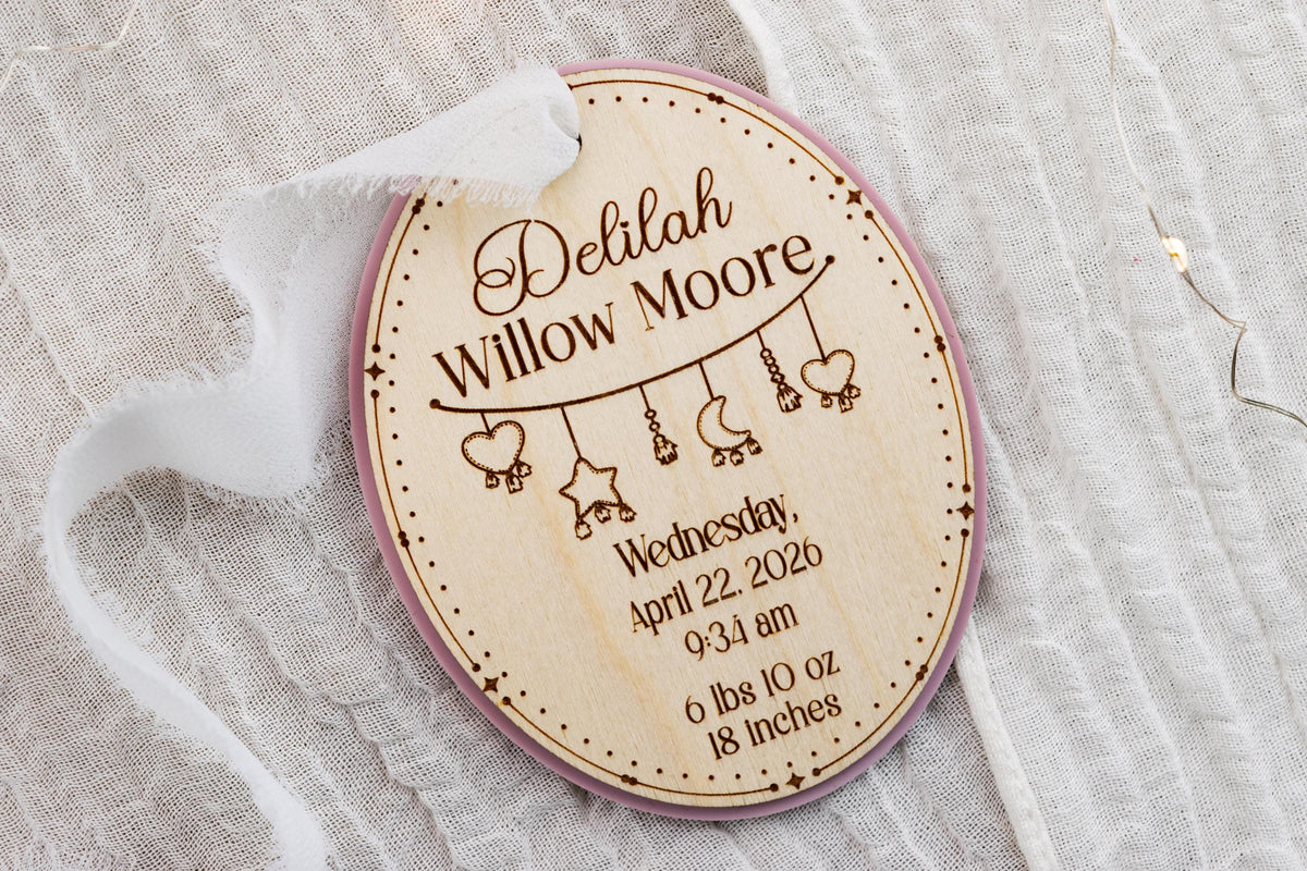 Angled view of an oval wooden baby keepsake ornament with pink acrylic backing, engraved birth details, and white ribbon.