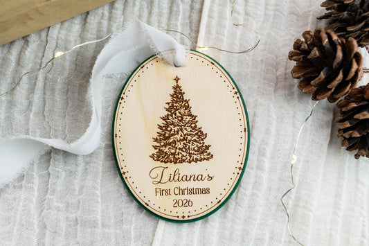 Oval wooden Baby's First Christmas ornament with engraved vintage Christmas tree, Liliana's name, 2026, and green acrylic backing.