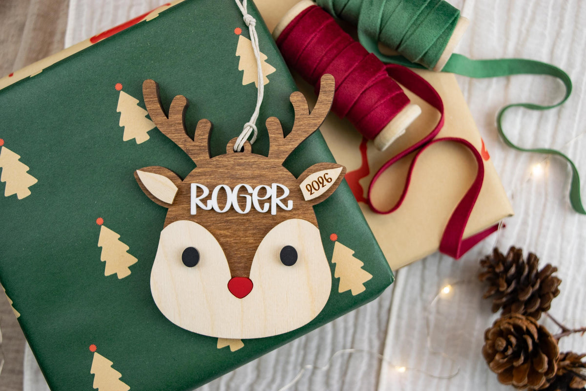Personalized layered wood reindeer ornament with brown antlers, red nose, and raised white name Roger, displayed on Christmas gift wrap.